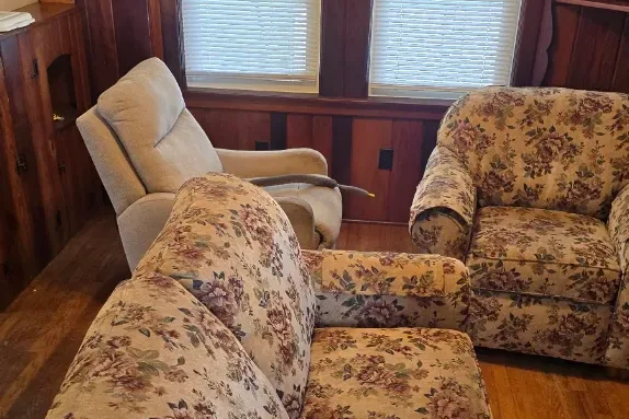 A wood-paneled living room filled with a floral couch, matching armchair and a recliner, blocking the floor.