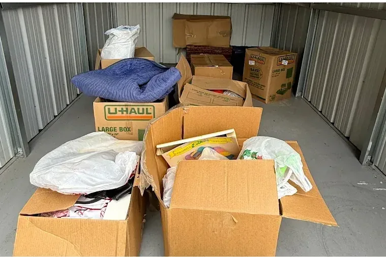 A storage unit with boxes, bags and bedding piled on the floor.